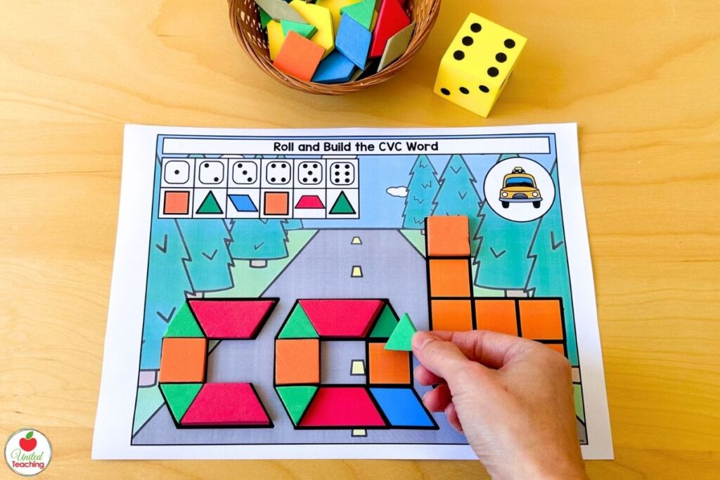 Hand placing a green triangle on a roll and build CVC word pattern block mat with a transportation theme, where students roll a die to choose which shape to place next